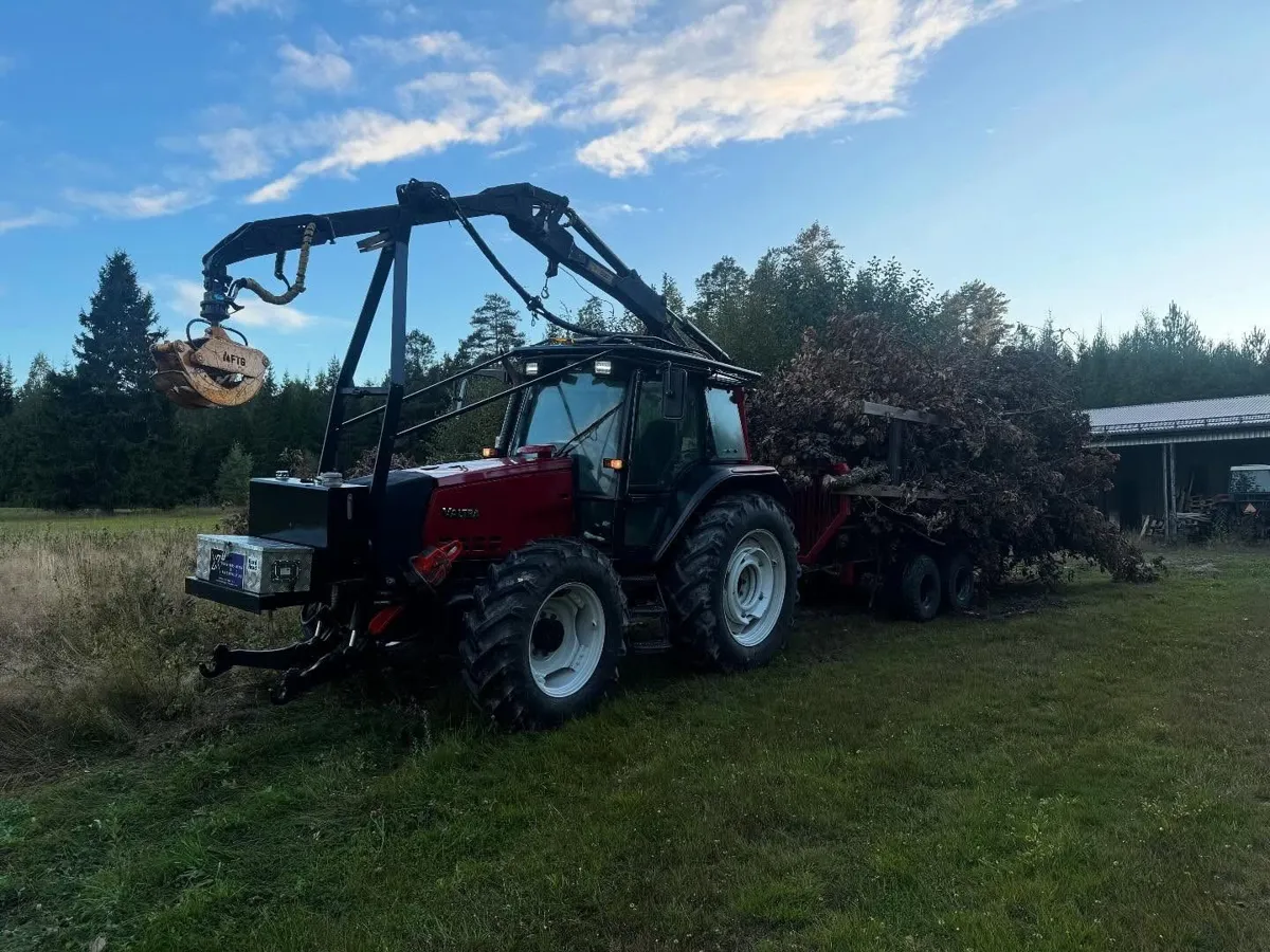 Röd traktor med skogskran och gripklo drar ett lass sly över en gräsmatta, granskog och blå himmel bakom