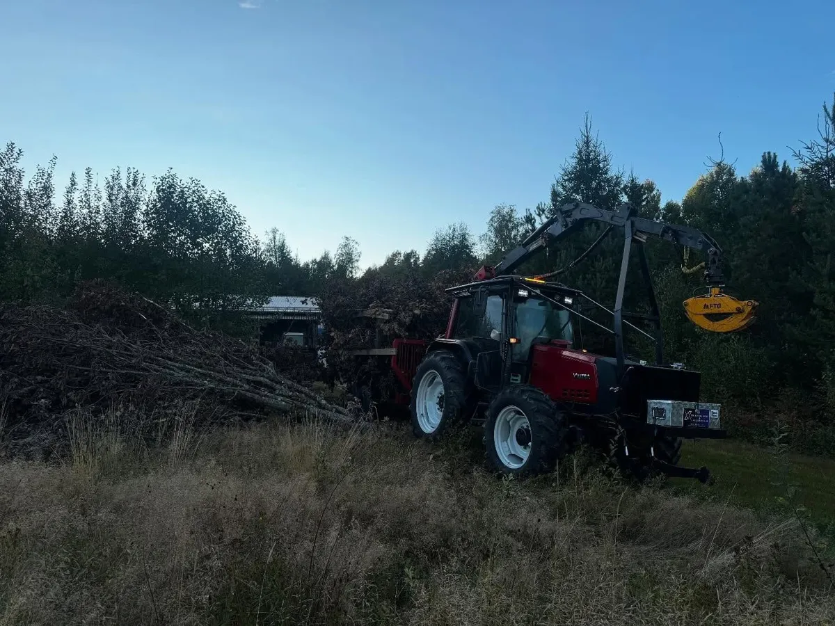 Traktor med kran drar ett lass sly genom högt gräs i skymningen