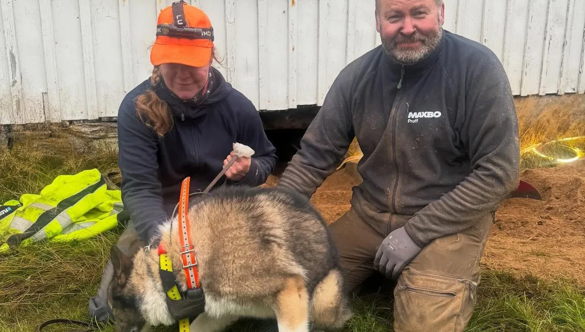 Två personer på knä i gräset med en hund i sele framför ett vitt hus