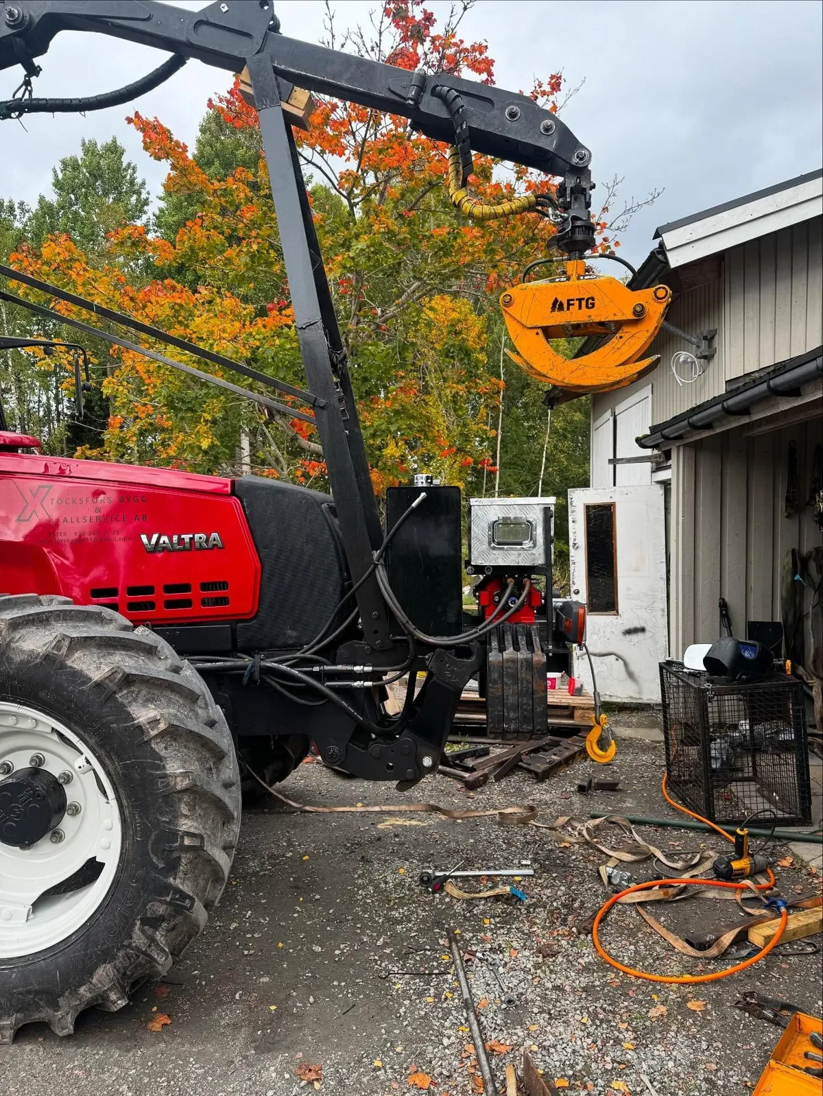 Skogskran med orange gripklo över en röd traktor, höstfärgad lönn i bakgrunden